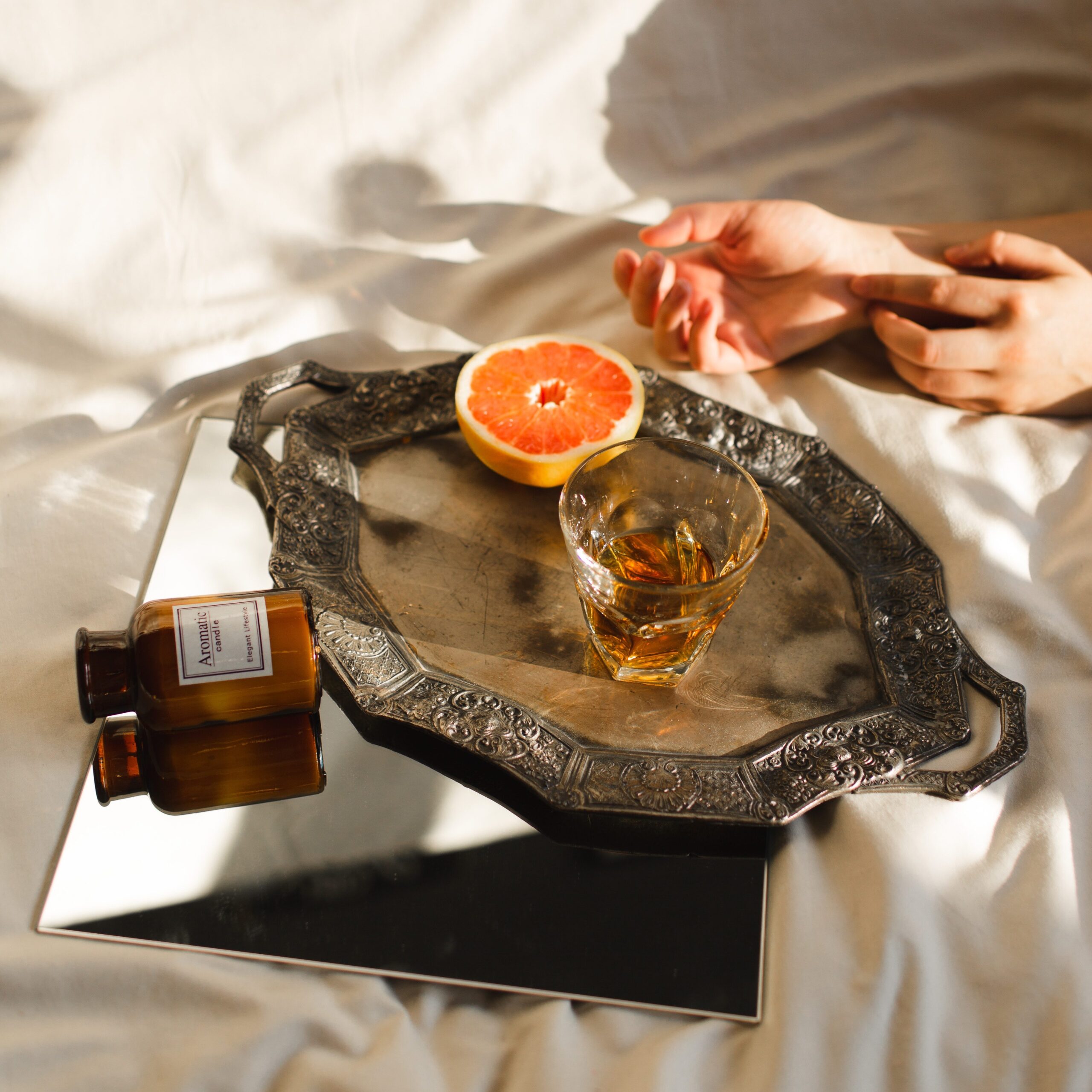 An alcoholic drink in a glass next to a half orange put on a tray that's on a mirror with an empty medicine bottle, all on bed sheets next to two hands.